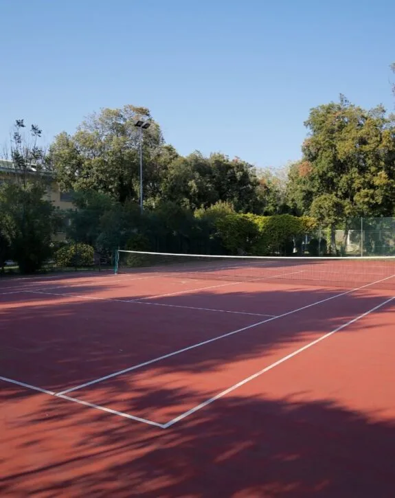 Campo da tennis all'aperto con superficie rossa, rete centrale e alberi verdi sullo sfondo sotto cielo sereno.