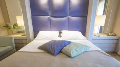 Letto matrimoniale con cuscini colorati, testiera imbottita viola e lampade accese sui comodini laterali.