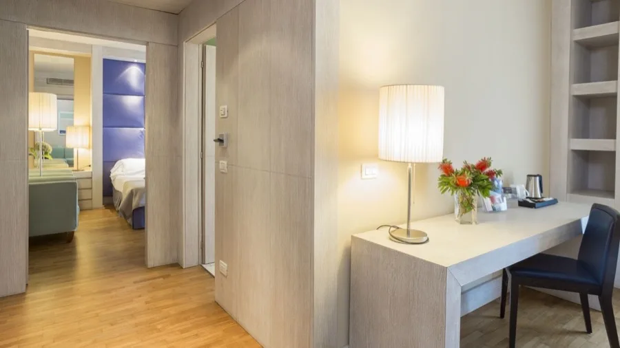 Interno di una stanza con pavimento in legno, scrivania con lampada e fiori, e letto visibile in una stanza adiacente.