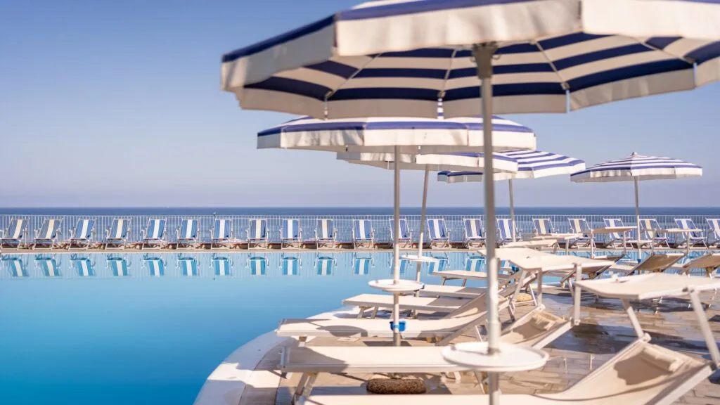 Piscina con lettini bianchi e ombrelloni a righe blu e bianche, vista sul mare e cielo sereno.