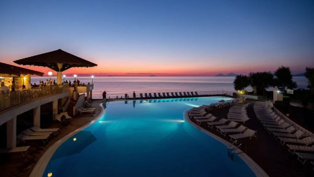 Piscina illuminata di un hotel al tramonto, con sdraio vuote e vista sul mare calmo all'orizzonte.