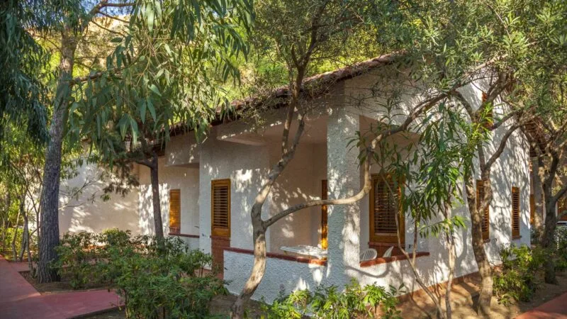 Casa bianca con tetto in tegole e persiane in legno, circondata da alberi e piante verdi in un giardino soleggiato.