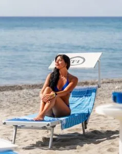 Donna in bikini blu seduta su lettino con asciugamano blu sulla spiaggia al sole, mare sullo sfondo.