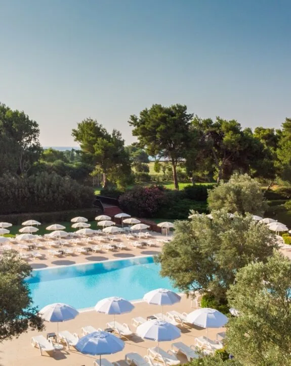 Piscina all'aperto con ombrelloni e sdraio bianchi, circondata da alberi verdi e cielo sereno.