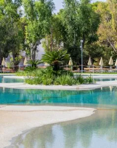 Un tranquillo giardino con piscine blu e vegetazione, circondato da alberi e ombrelloni beige.