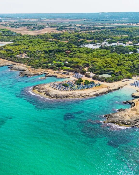 Vista aerea di una spiaggia con ombrelloni blu e mare turchese, circondata da una vegetazione verdeggiante.