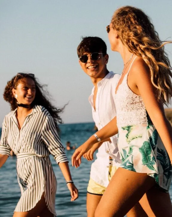Tre giovani sorridenti con occhiali da sole camminano sulla spiaggia al tramonto, onde sullo sfondo.
