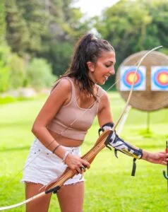 Donna sorridente con arco e protezione per il braccio mentre pratica tiro con l'arco all'aperto davanti a due bersagli colorati.