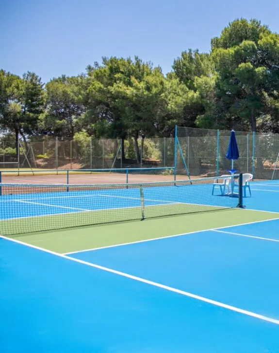 Campo da tennis vuoto con rete, sedie bianche e ombrellone blu, circondato da alberi verdi.