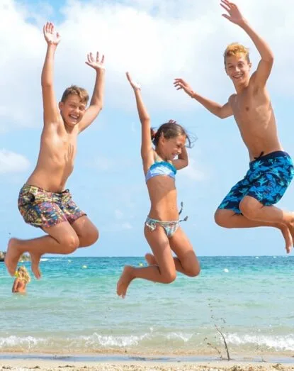 Tre bambini sorridenti saltano su una spiaggia con il mare e il cielo azzurro sullo sfondo.