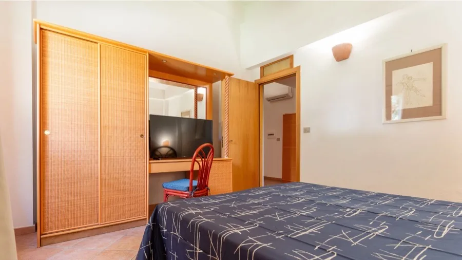 Camera da letto con letto matrimoniale, armadio in legno chiaro, poltrona rossa e TV su mobile con specchio.