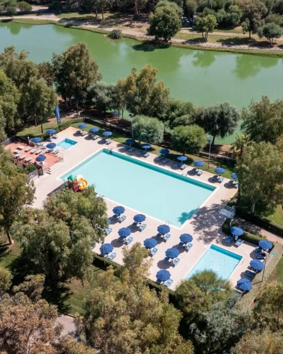 Piscina all’aperto con ombrelloni blu e sdraio, circondata da alberi, vicino a un lago verde.