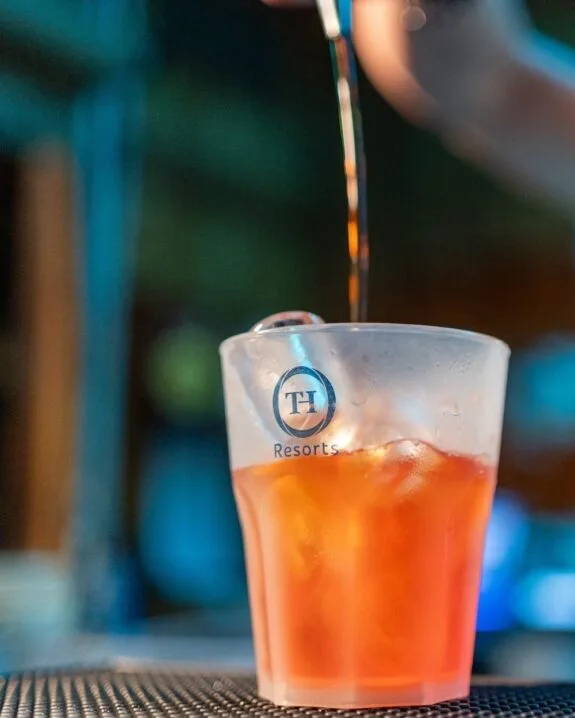 Bicchiere di plastica con logo TH Resorts contenente cocktail arancione con ghiaccio, mentre viene versato un liquido.