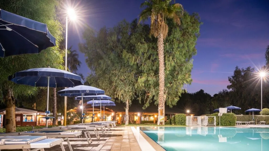 Piscina all'aperto illuminata di sera con sdraio e ombrelloni blu, circondata da alberi e palme.