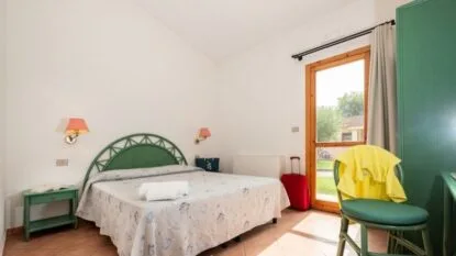 Camera da letto luminosa con letto matrimoniale, comodino verde, valigia rossa e sedia con indumento giallo vicino a porta finestra.