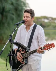 Uomo che suona la chitarra e canta al microfono all’aperto, indossa camicia bianca, sfondo naturale con alberi e lago.