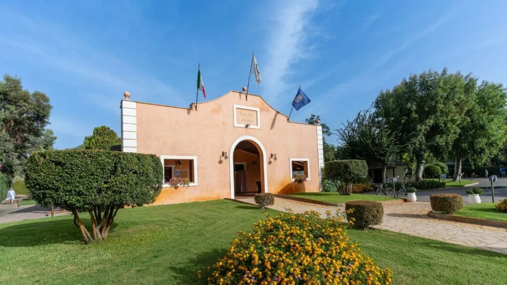 Ingresso di un edificio color pesca con tre bandiere, circondato da prato verde e siepi potate.