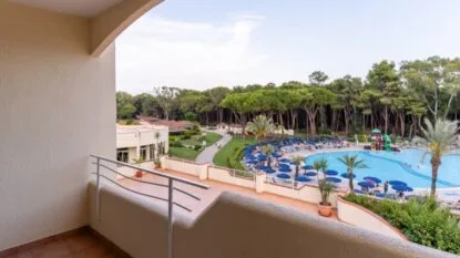 Vista di una piscina all'aperto con ombrelloni blu, circondata da alberi, ripresa da un balcone con ringhiera bianca.