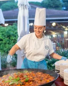 Cuoca sorridente con cappello bianco e grembiule azzurro mentre cucina in una grande padella all'aperto.