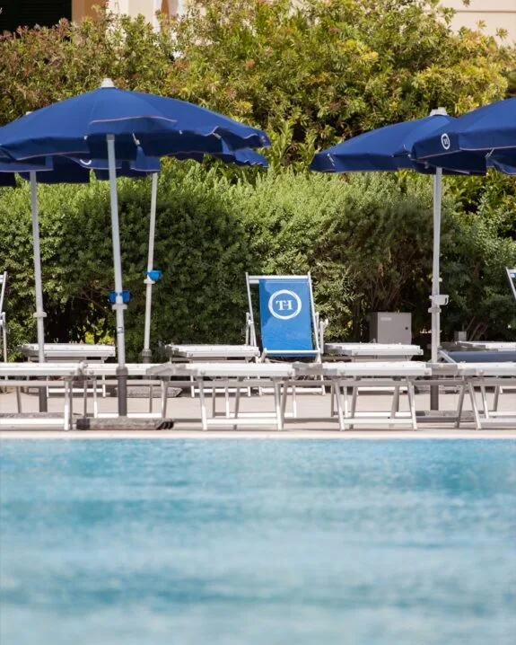 Piscina con lettini e ombrelloni blu, sfondo di cespugli verdi e prato soleggiato.