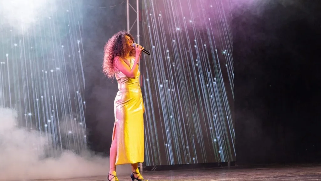 Cantante donna con abito lungo dorato e capelli ricci su palco con microfono e sfondo luminoso astratto.