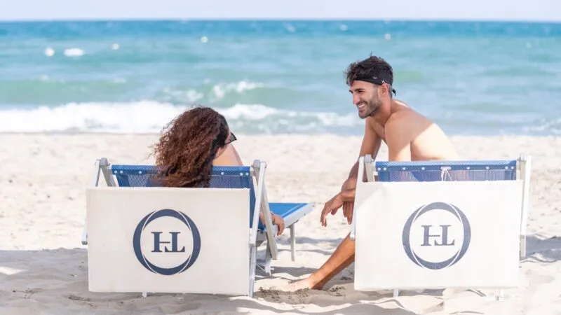 Coppia seduta su due sedie da spiaggia con logo HL, che chiacchiera sorridendo davanti al mare calmo.