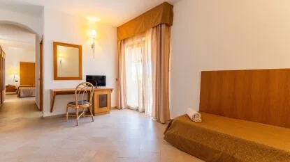 Camera d'albergo luminosa con letto singolo, scrivania, sedia e balcone con tende color crema e beige.
