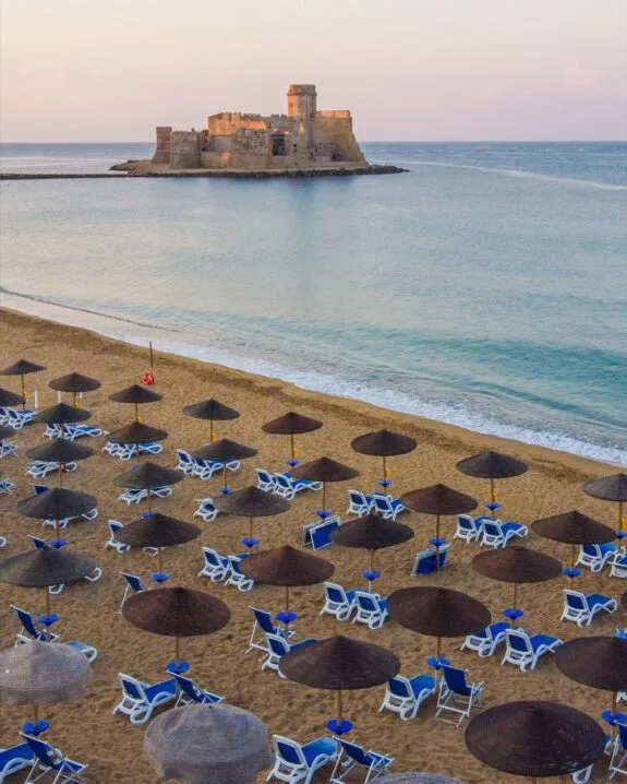 Spiaggia con lettini e ombrelloni, castello antico su isola rocciosa nel mare calmo al tramonto.