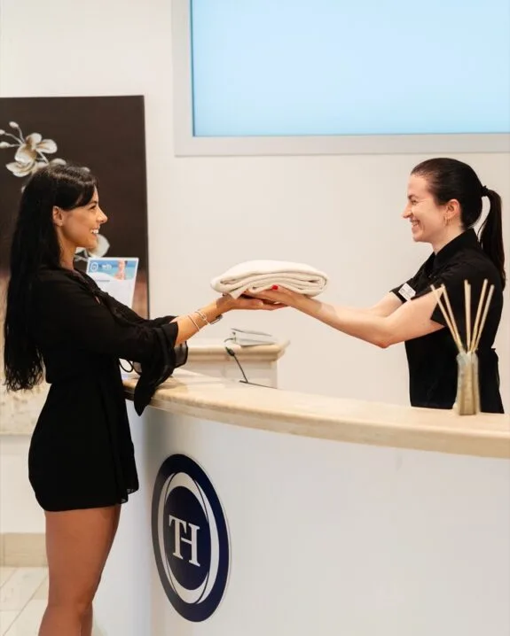 Due donne sorridenti si scambiano un asciugamano piegato al banco di accoglienza di un centro benessere.