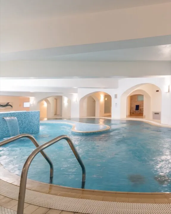 Piscina coperta con acqua azzurra, scaletta in metallo e cascatella, illuminazione soffusa e archi bianchi.