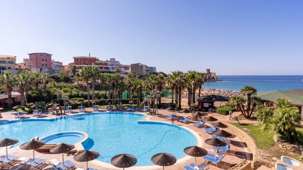 Piscina con ombrelloni e lettini, palme, spiaggia affollata e mare calmo con castello sullo sfondo.