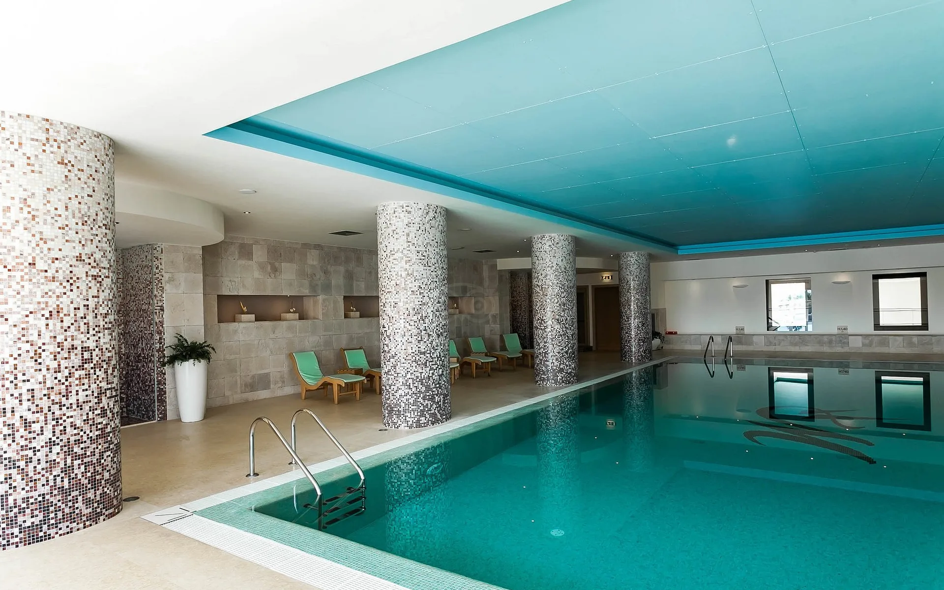 Piscina coperta con colonne rivestite a mosaico, lettini e soffitto azzurro