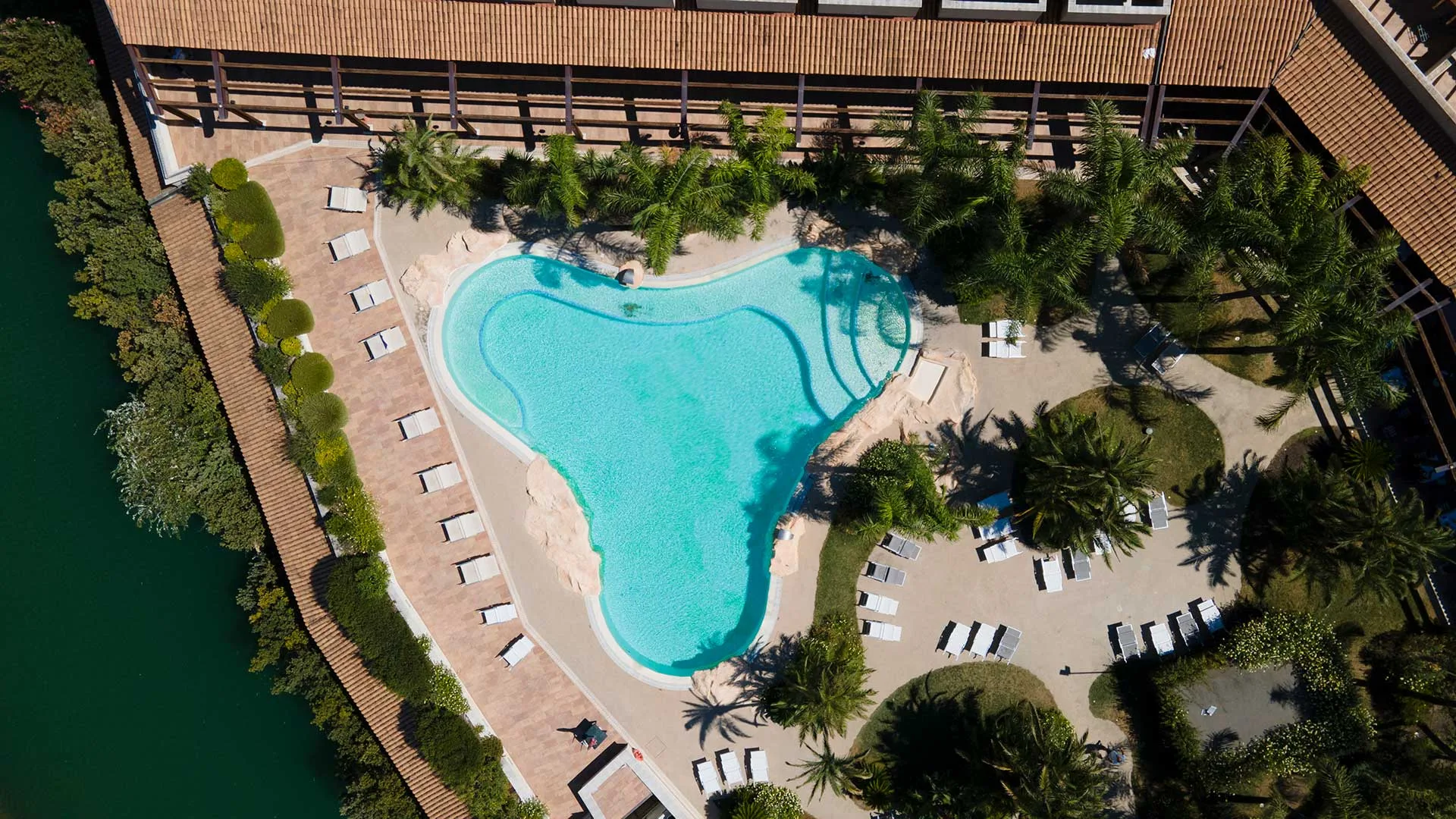 Vista aerea dall'alto della piscina esterna circondata da palme e lettini del resort