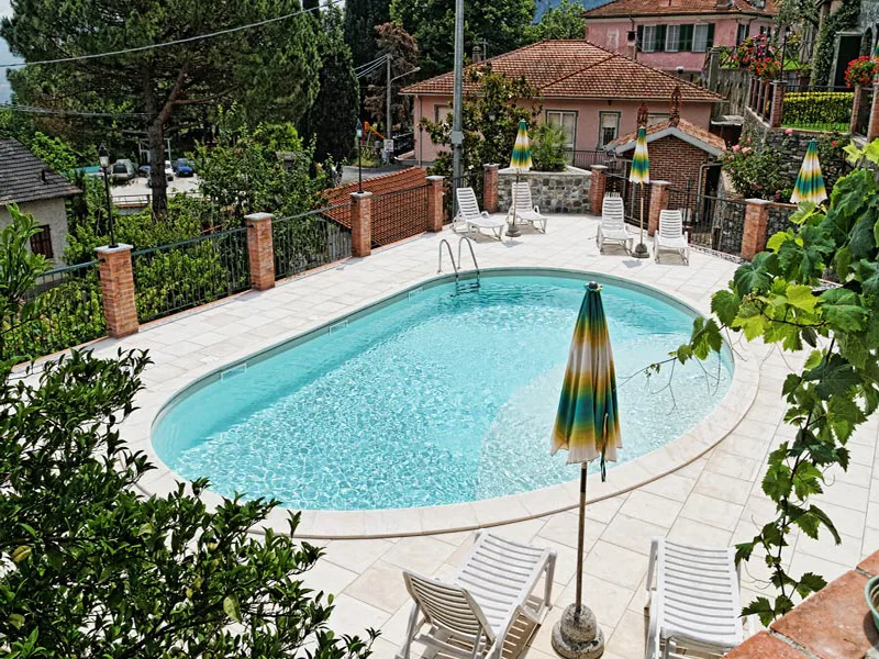 Piscina ovale con lettini e ombrelloni circondata da vegetazione e case del borgo