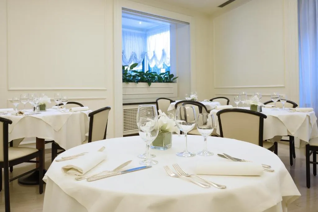 Sala ristorante con tavoli rotondi apparecchiati, tovaglie bianche e finestra con piante