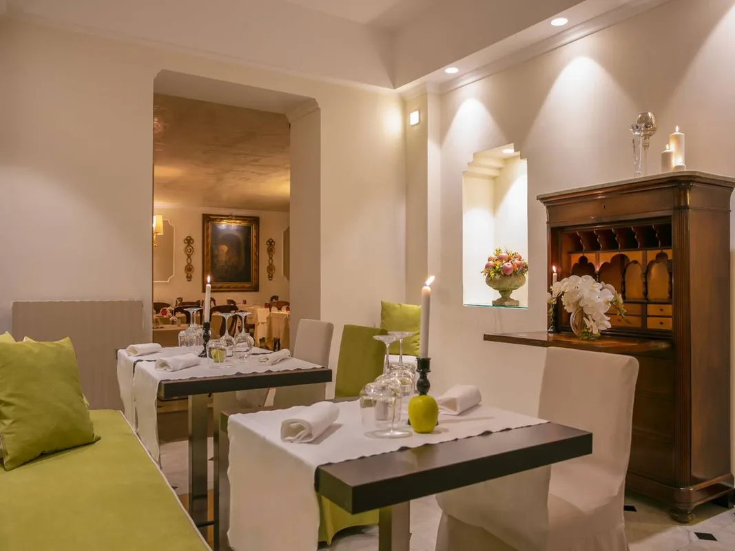 Saletta ristorante intima con tavoli apparecchiati, candele, mobile antico in legno e cuscini verdi
