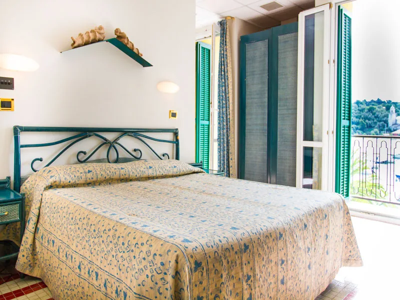 Camera matrimoniale con letto in ferro battuto, persiane verdi e balcone con vista sul porto
