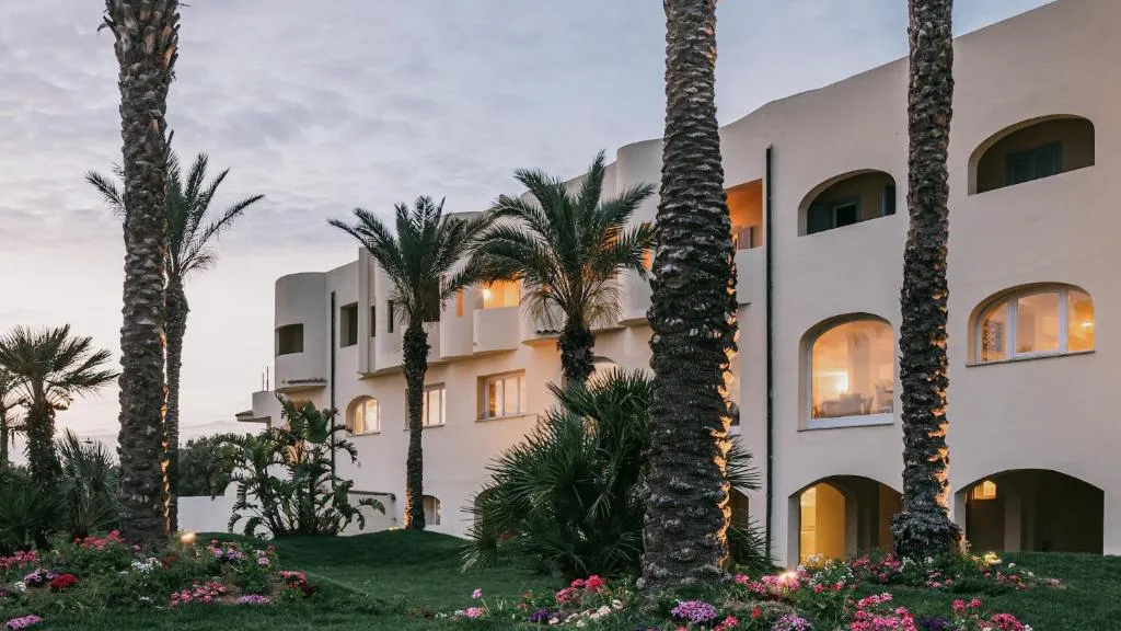 Facciata del resort con archi illuminati, palme e aiuole fiorite al tramonto