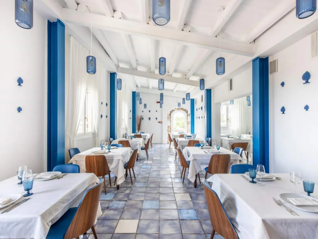 Sala ristorante luminosa in bianco e blu con tavoli apparecchiati, lampade pendenti e decorazioni in ceramica siciliana