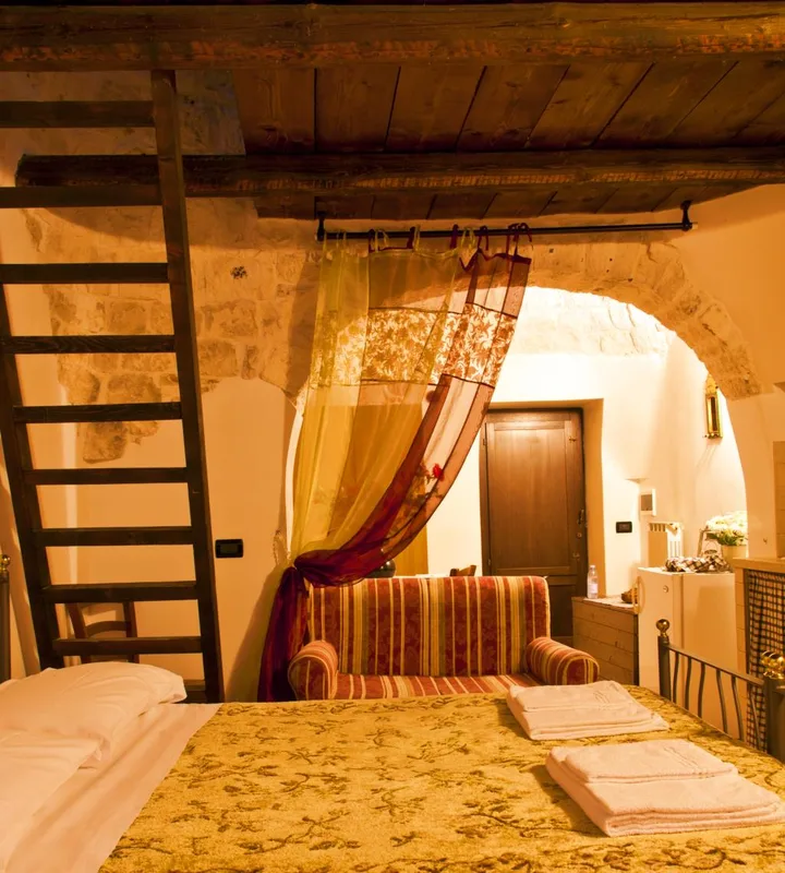 Camera in trullo con illuminazione calda, letto, divanetto e scala in legno