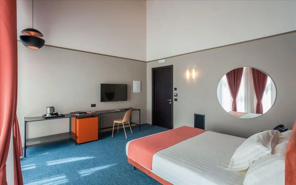 Camera moderna con moquette blu, letto matrimoniale, specchio rotondo e scrivania con TV