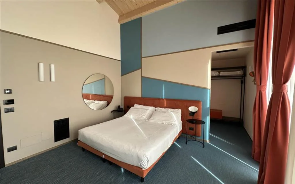 Camera con parete geometrica azzurra, letto matrimoniale, specchio rotondo e angolo guardaroba