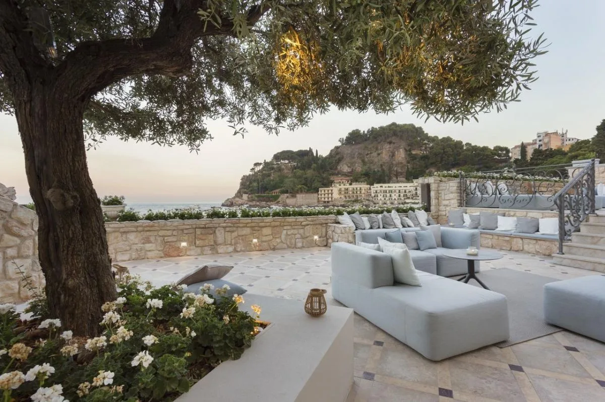 Terrazza panoramica con divani chiari sotto ulivo secolare vista costa al tramonto