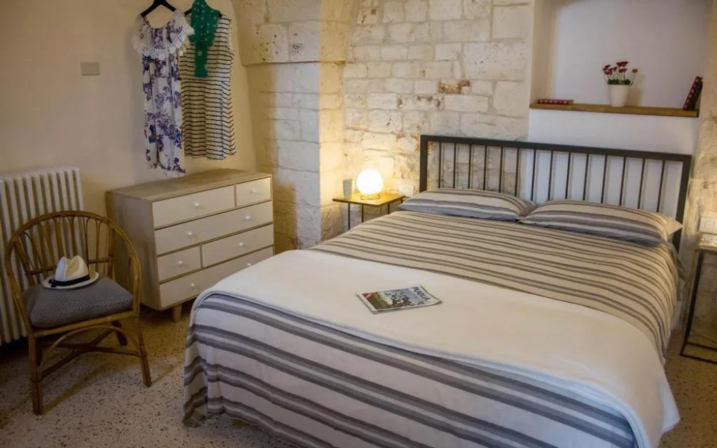 Camera con letto a righe, cassettiera in legno chiaro, pareti in pietra e abiti appesi