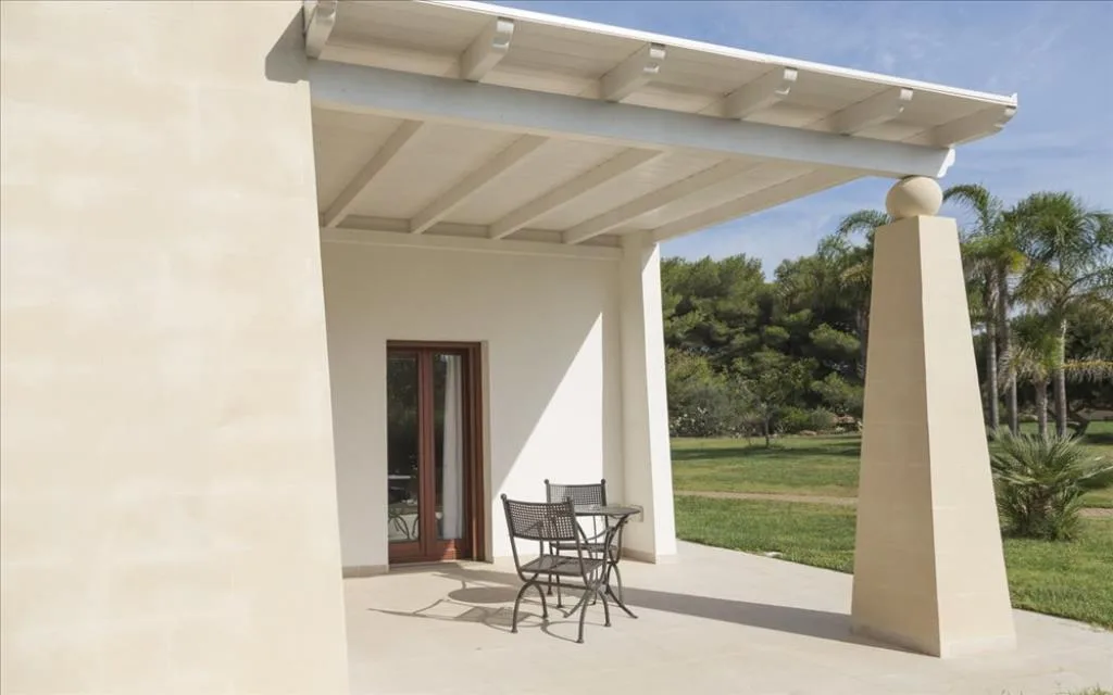 Veranda coperta con pergolato in legno, tavolino e sedie in ferro affacciata sul giardino con palme