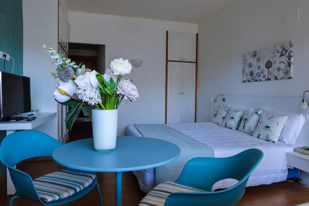 Camera con letto matrimoniale, tavolino azzurro con fiori, sedie e angolo TV