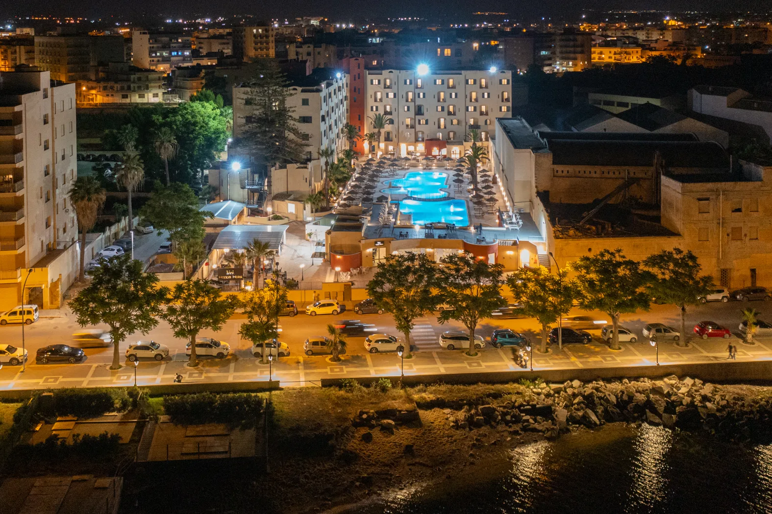 Vista notturna aerea dell'hotel con piscine illuminate, lungomare alberato e luci della città