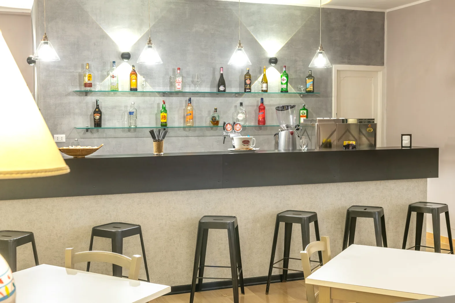 Bancone bar dell'hotel con bottiglie di liquori su mensole in vetro, sgabelli neri e lampade a sospensione