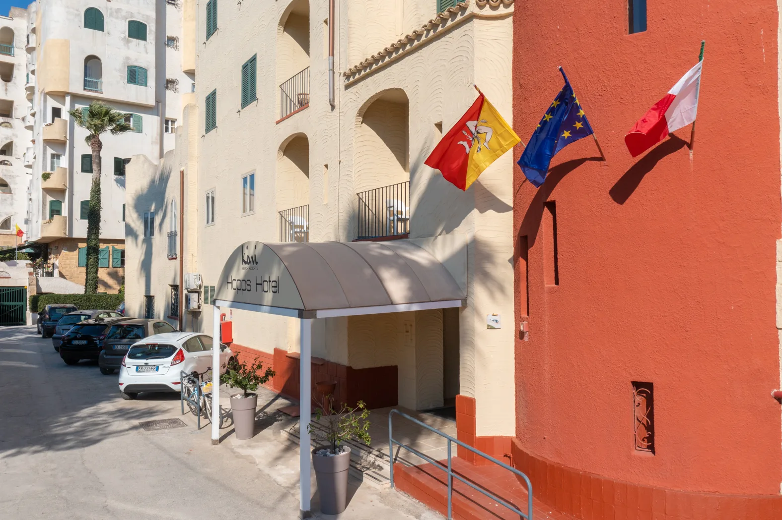Ingresso dell'Hopps Hotel con pensilina, bandiere di Sicilia, Europa e Italia e facciata color panna e rosso