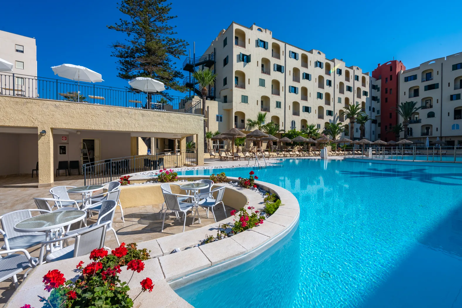 Piscina blu con bordo curvo, tavolini e sedie bianche, fiori rossi e vista sull'edificio dell'hotel